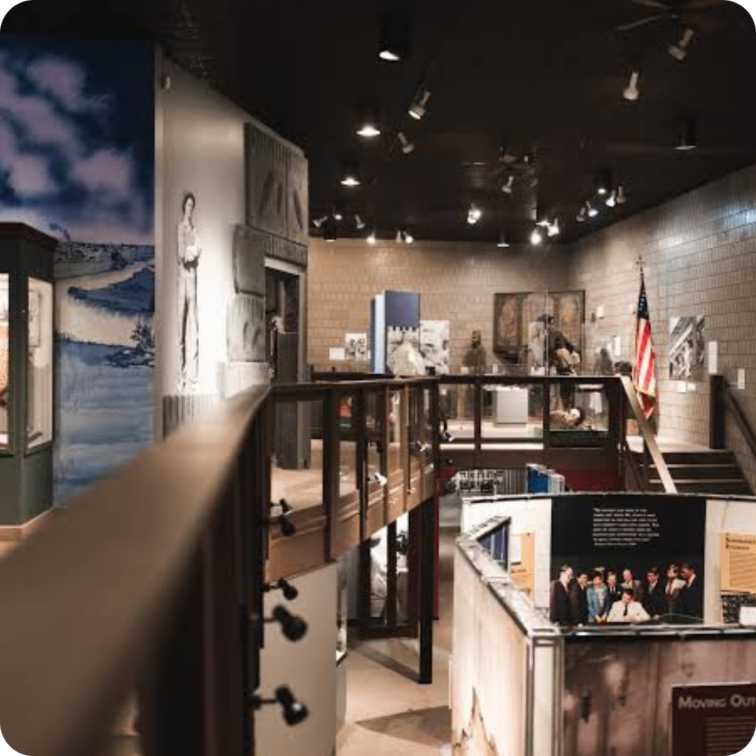 A museum interior with exhibits, informational displays, photographs, and an American flag, viewed from an elevated walkway under ceiling spotlights.
